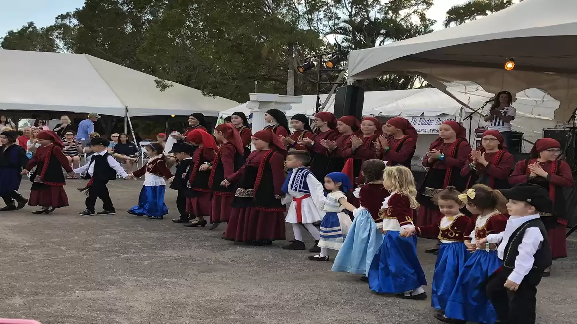 Children and adults in traditional costumes dancing at an outdoor performance in front of an audience.