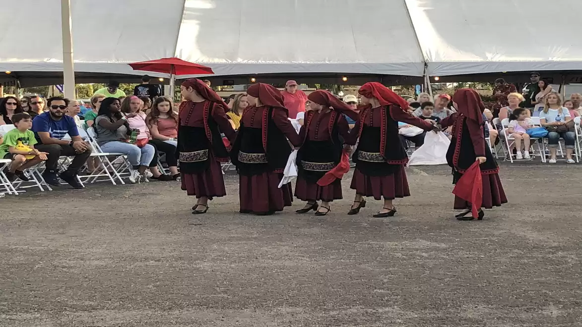 Traditional dancers in red attire perform a folk dance at a festival, surrounded by an audience seated on white chairs.