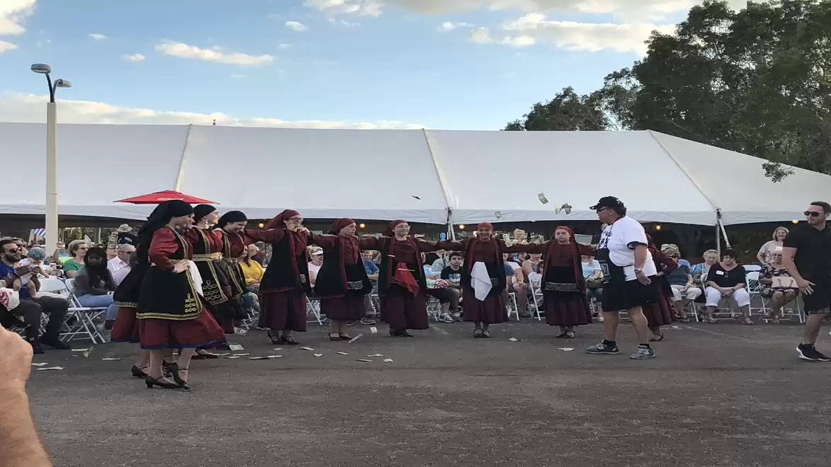 A group of people in traditional clothing dancing at an outdoor event. Spectators are seated on chairs around them. In the background, there is a white tent and trees.