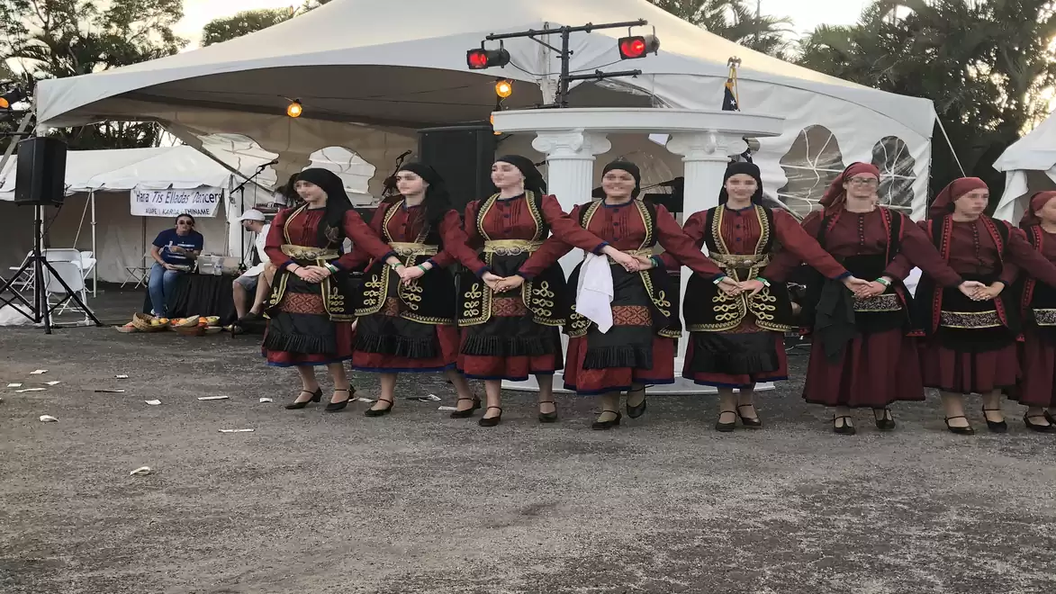 Group of women in traditional costumes standing on a square in front of white tents, surrounded by lighting and trees.