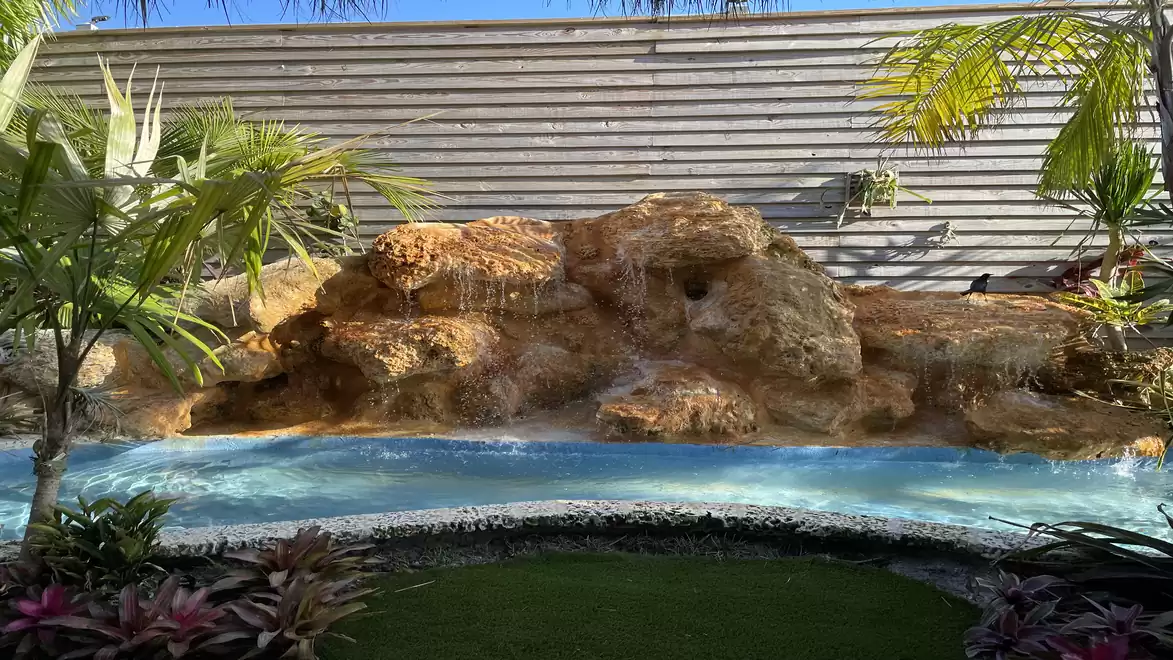 Image of an artificial waterfall with shallow water and surrounding tropical vegetation.