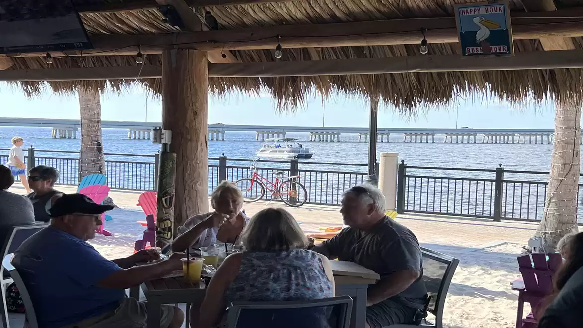 People sitting under a thatched roof at a table in a beach café with a view of the sea and a bridge in the background.