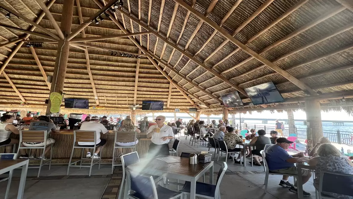 A lively beachside restaurant with a thatched roof, filled tables, and guests enjoying the food and view.
