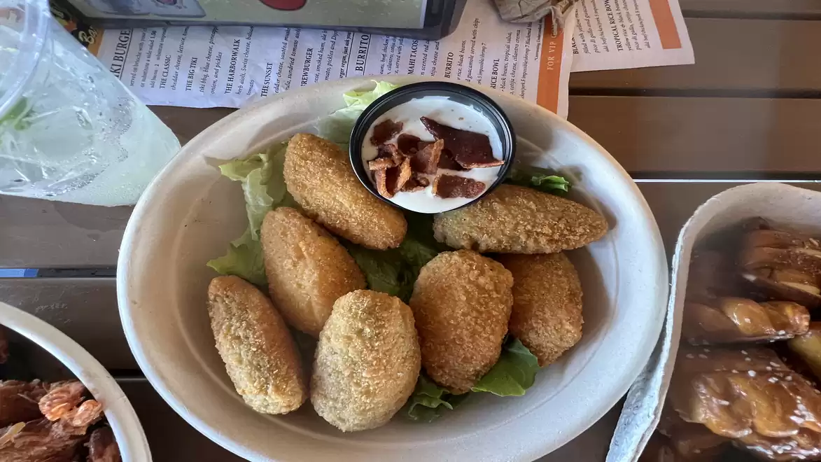 A plate of breaded snacks next to a dip with bacon pieces, served with a drink in the background.
