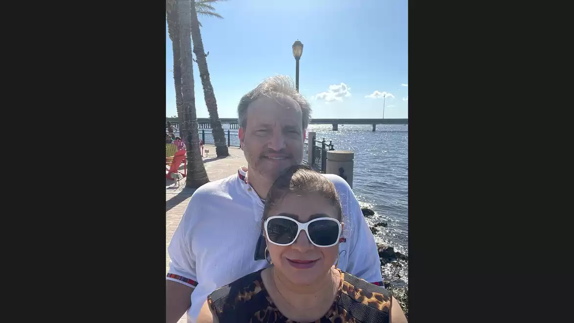 A man and a woman smile at the camera while standing on a sunny day at a waterfront. Palm trees and the sea are visible in the background.