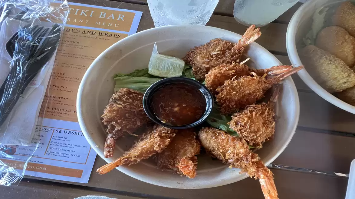 Bowl of crispy fried shrimp with a dip, green beans, and lime wedge, with a menu and drink in the background.