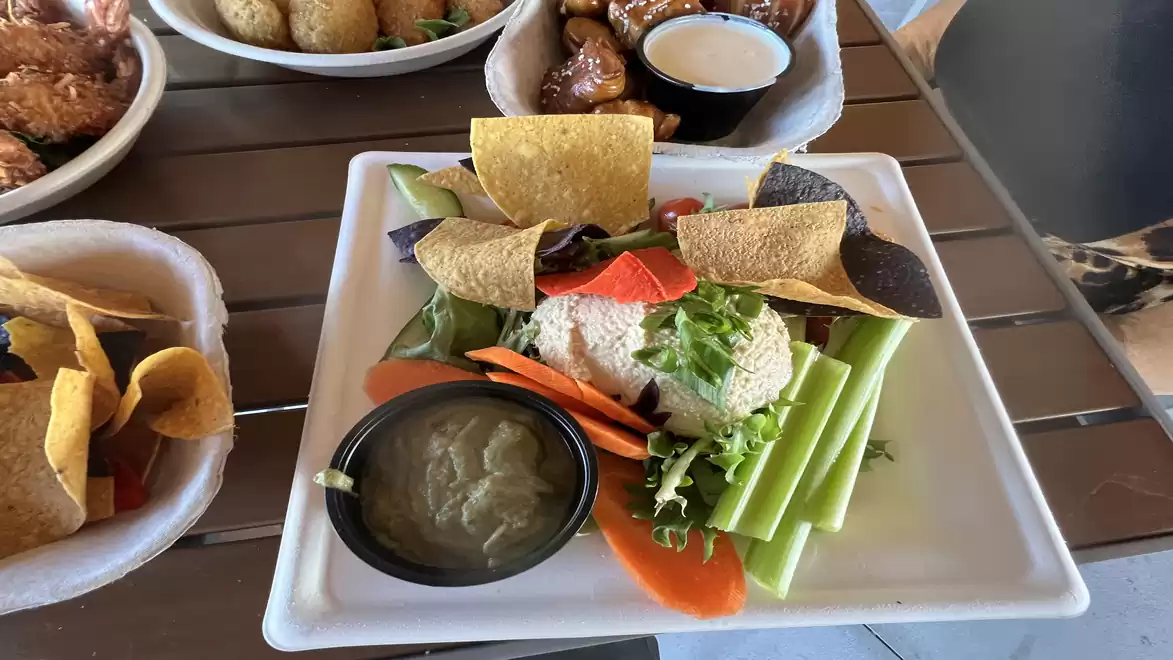A plate with fresh vegetables including celery sticks, bell peppers, and carrots, accompanied by a portion of cottage cheese and tortilla chips. In the background, there are other bowls with various dishes.