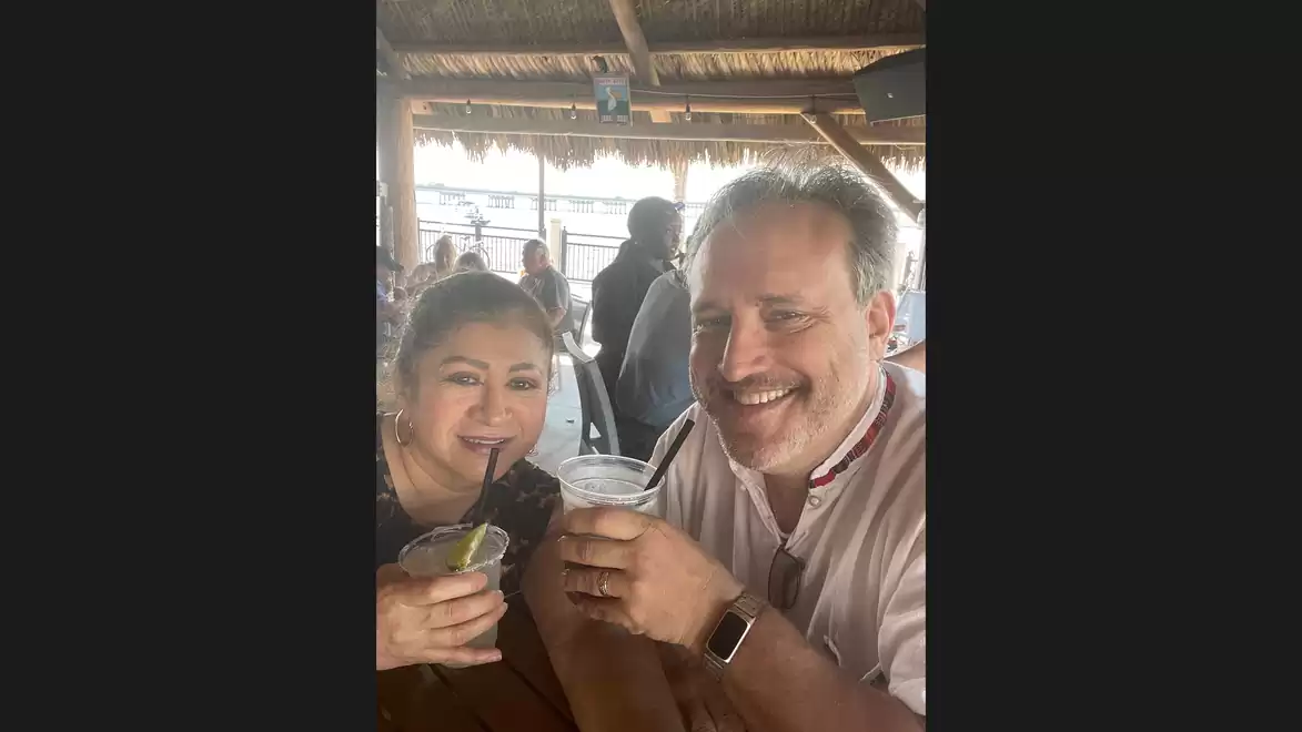 A man and a woman smiling and each holding a drink. They are sitting in a restaurant with a wooden roof and other guests in the background.