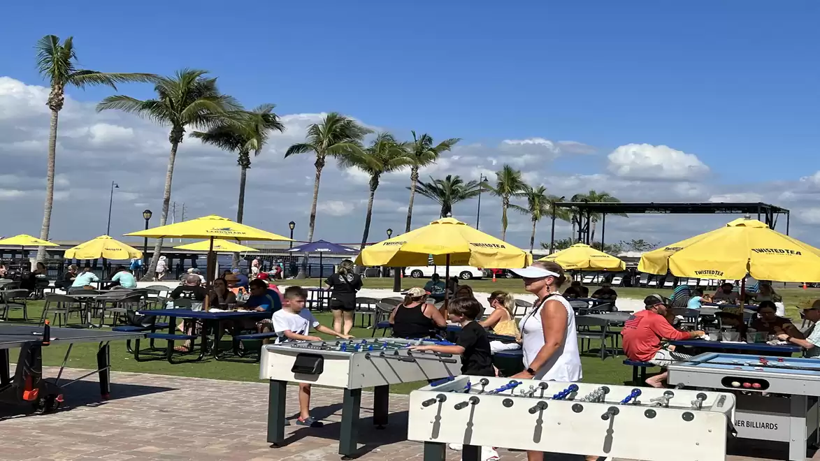 An outdoor terrace with several people at foosball tables and yellow umbrellas, surrounded by palm trees and a clear sky.
