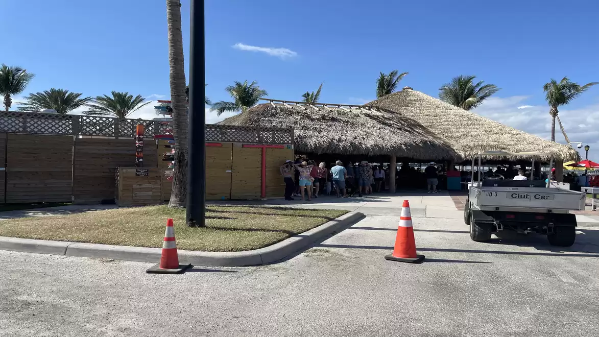 Image of a pavilion with a thatched roof, groups of people underneath. There are palm trees and orange traffic cones on the side.