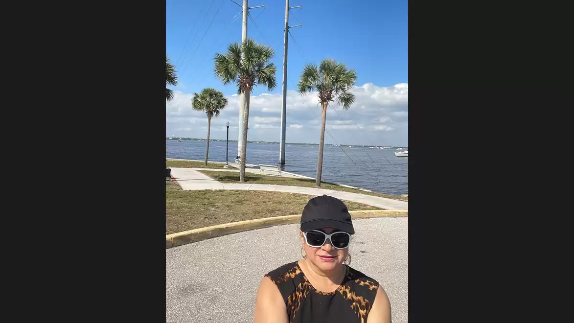 Person wearing sunglasses and a cap stands in front of a coastal landscape with palm trees and a blue sky.