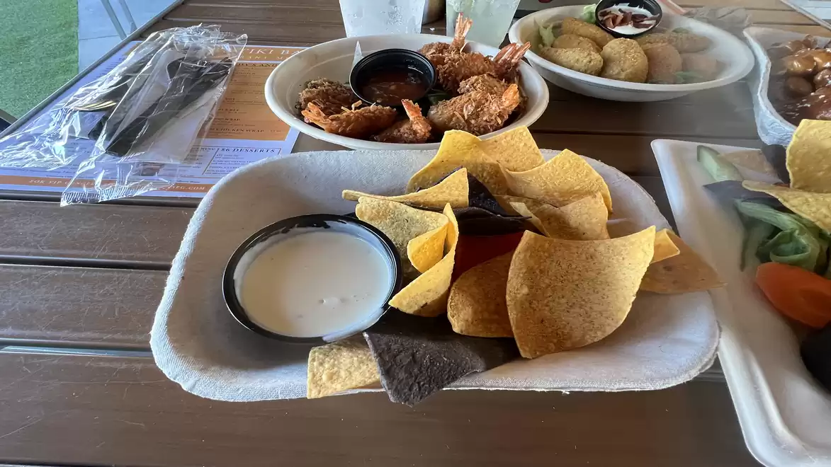 A table with a plate of nachos and dip on a wooden table, behind it a plate of fried shrimp and another plate with appetizers.