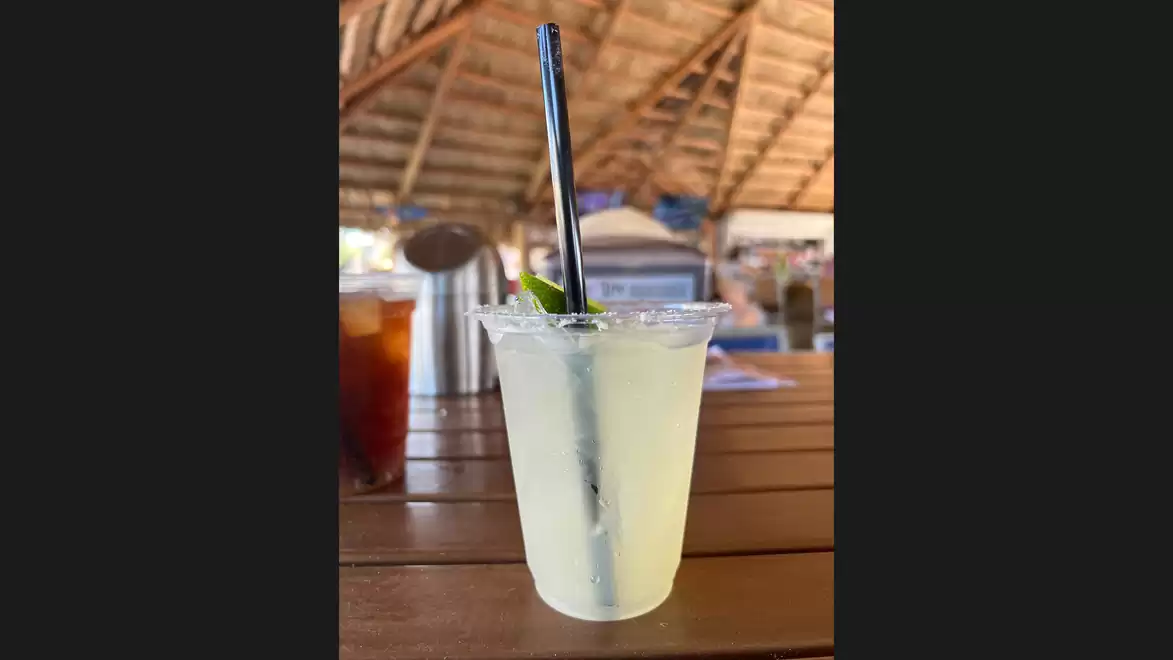 A refreshing drink in a plastic cup with a black straw and a lime wedge on a wooden table under a thatched roof.