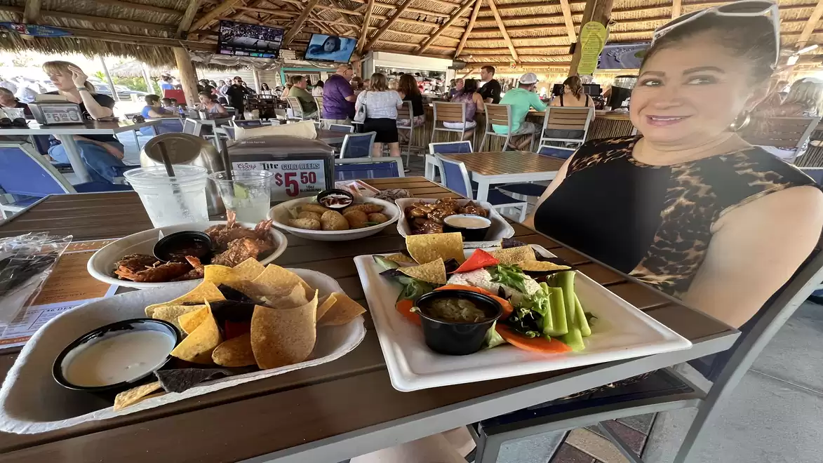 A restaurant with a rustic thatched roof. Various dishes, including salad, nachos, and dips, are on the table. People are sitting and chatting in the background.