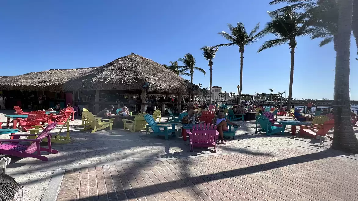 A sunny beach bar with a thatched roof and colorful lounge chairs, surrounded by palm trees under a blue sky.