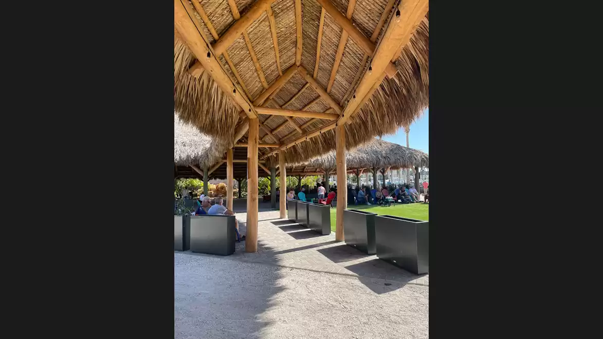 People sitting at tables under a thatched roof in a sunny outdoor area with green grass.