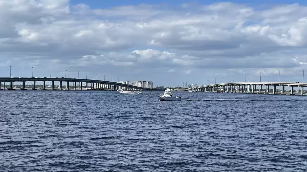 View of a wide waterway with two parallel bridges and a boat sailing in the middle. The sky is cloudy.
