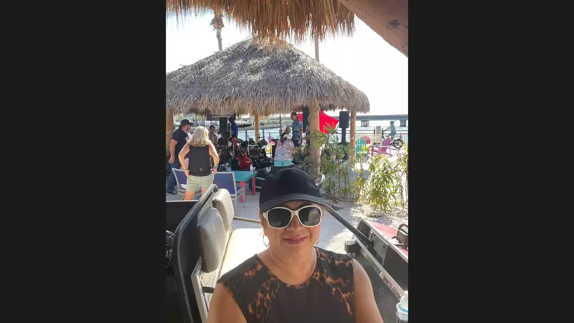Woman with sunglasses and hat sitting in a beach café, with people and the sea in the background.
