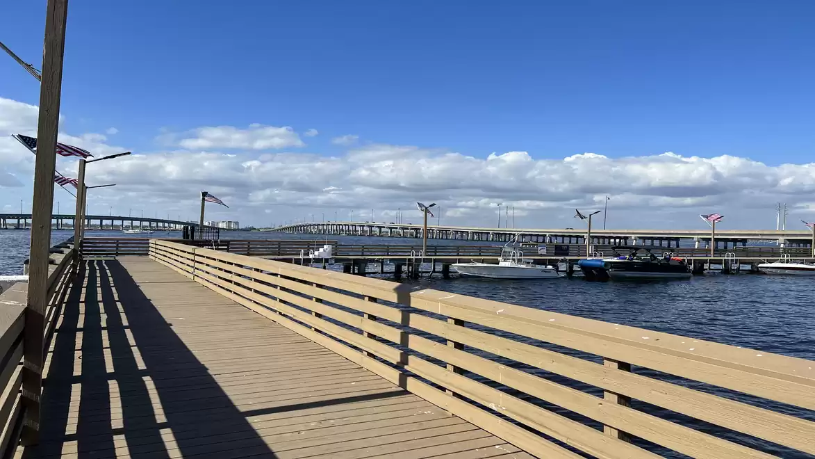 Wooden pier leading to a marina with several boats and a bridge in the background under a clear blue sky.