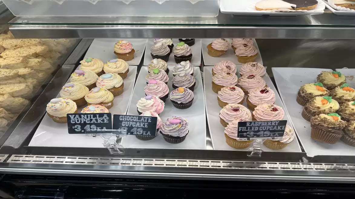 A glass display case filled with various cupcakes, including vanilla, chocolate, and raspberry cupcakes, with price tags.