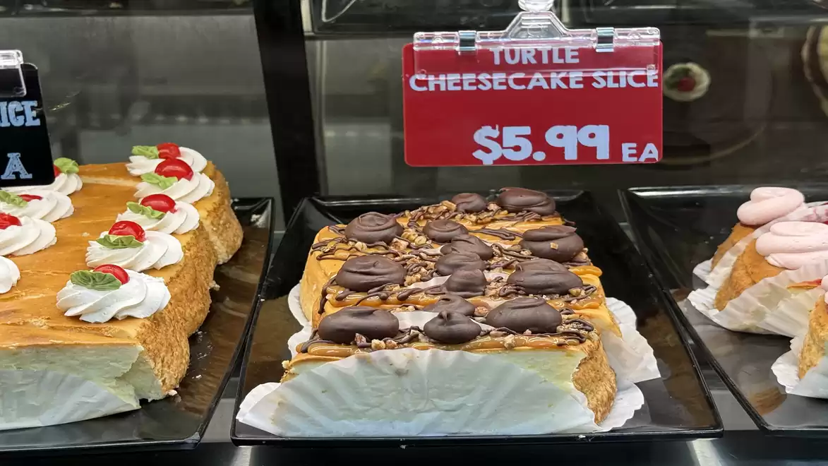 An image of three different cheesecake slices in a display, with a sign indicating a price of $5.99 for a Turtle Cheesecake Slice.