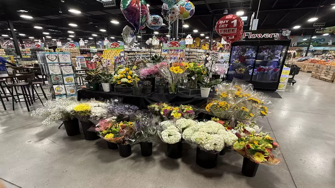 A flower stall in a large store displays a variety of colorful bouquets. In the background, balloons and other sales stands are visible.