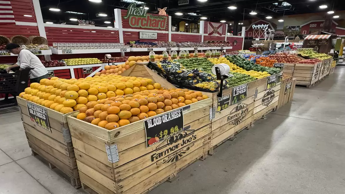 Interior view of a market with fresh fruits and vegetables stacked in wooden crates. A variety of fruits and vegetables can be seen, including oranges, apples, and avocados. The area is brightly lit, with several shelves visible in the background.