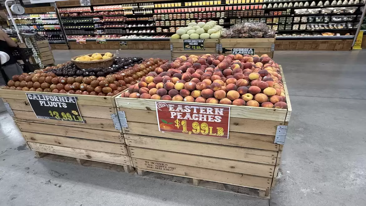Fresh fruit in wooden crates in a supermarket, with a sign displaying peaches and plums priced per pound.