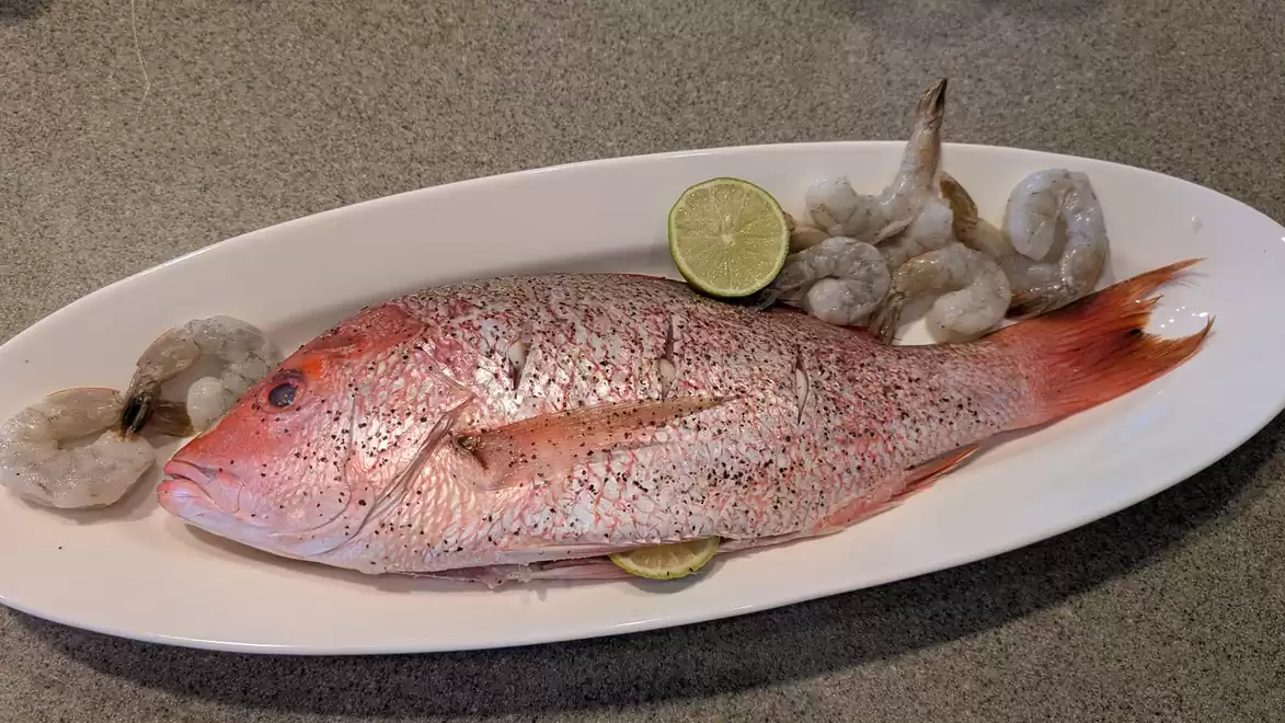 Fresh red fish - Red Snapper - and raw shrimp on an oval white plate with lime slices.