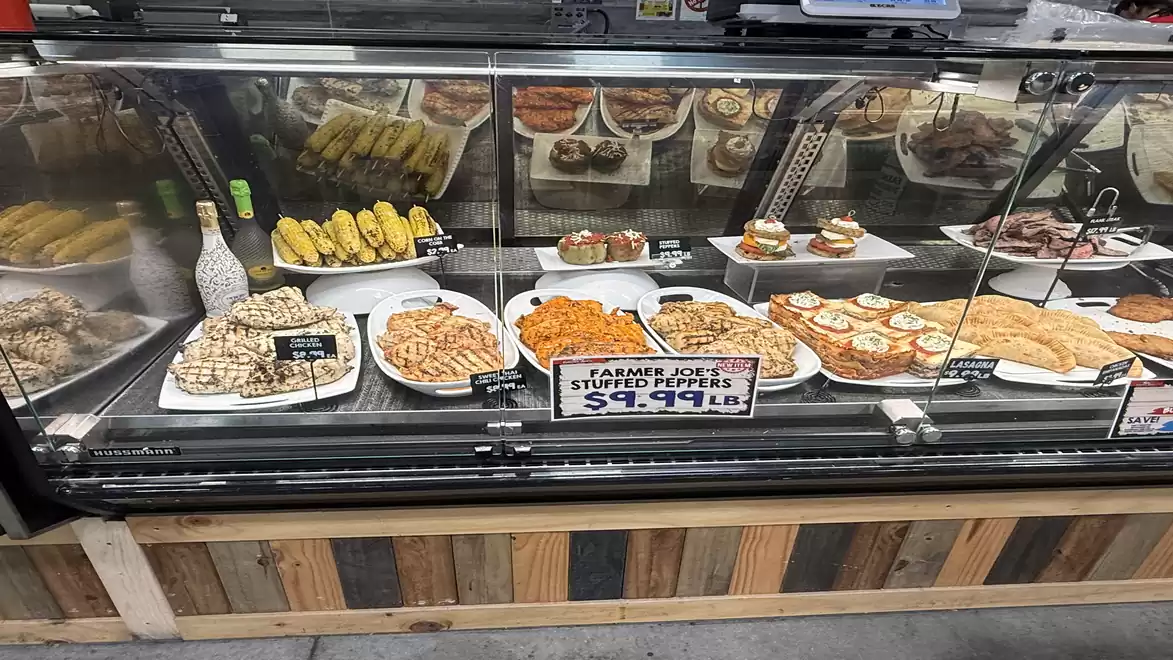 A refrigerated display case in a grocery store showcasing various dishes such as stuffed peppers, corn on the cob, and baked chicken pieces. A sign indicates the price of $9.99 per pound for stuffed peppers.