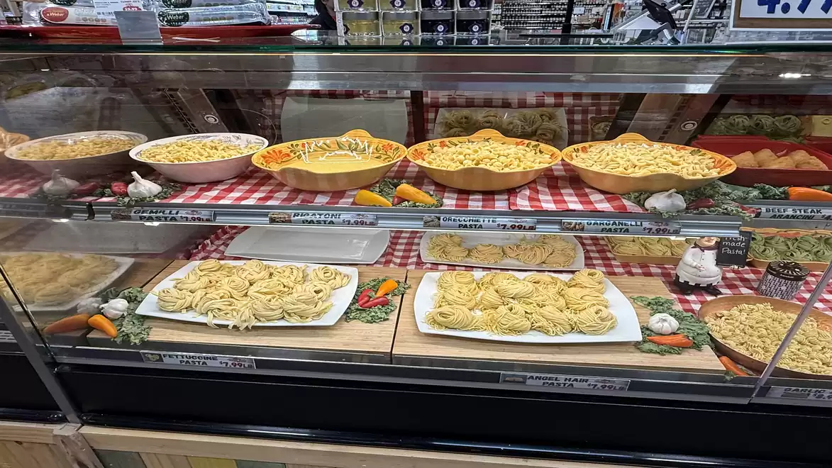 Display case with various pasta types in an Italian market hall. Bowls and plates with fresh pasta on red and white checkered tablecloths are visible.