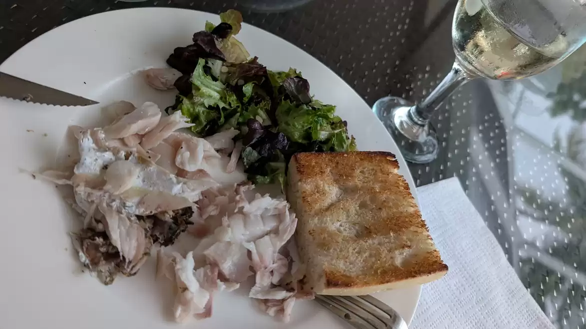 Plate with roasted fish, green salad, a slice of bread, and a glass of white wine on a table.