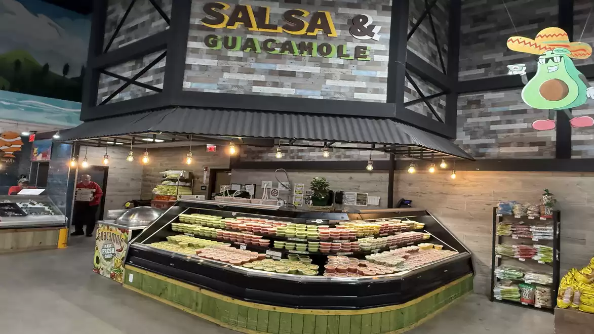 A store with a stand offering salsa and guacamole under a black roof structure. Various containers of dips are arranged on display. The store environment is modern and inviting.