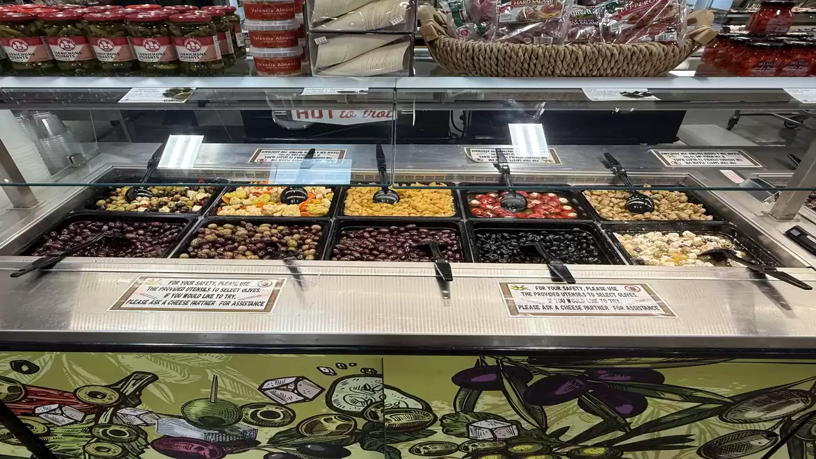 Close-up of a supermarket salad bar with various fresh ingredients in separate compartments, including olives, corn, and tomatoes.