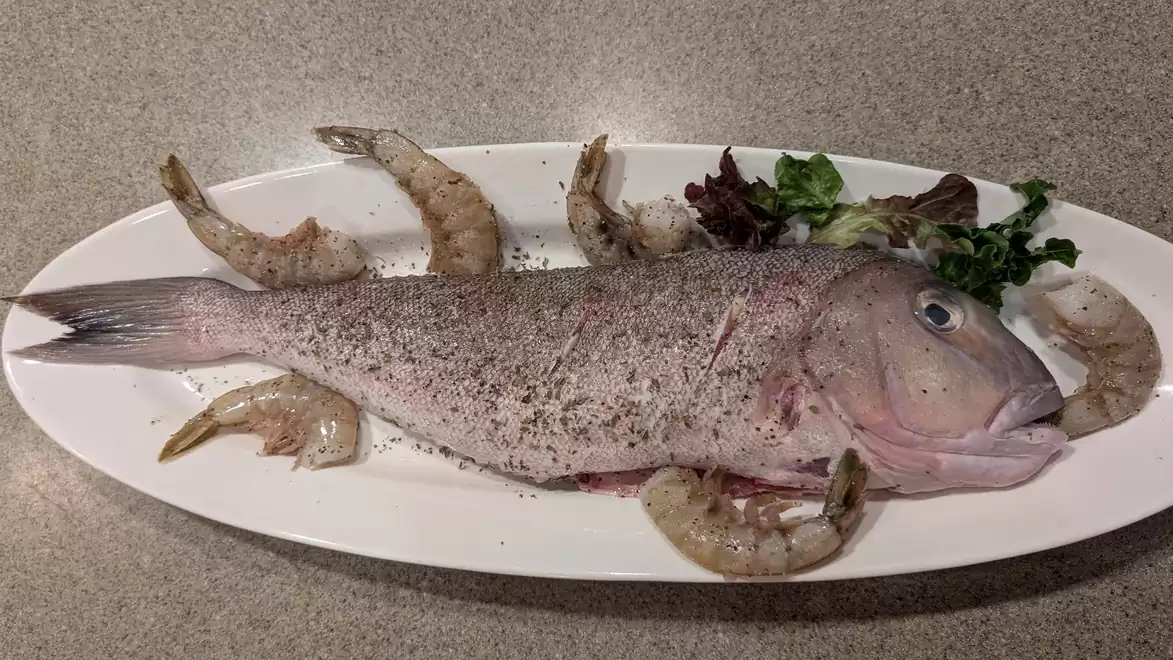 A whole fish, seasoned and served on a white plate, surrounded by shrimp and some green salad.