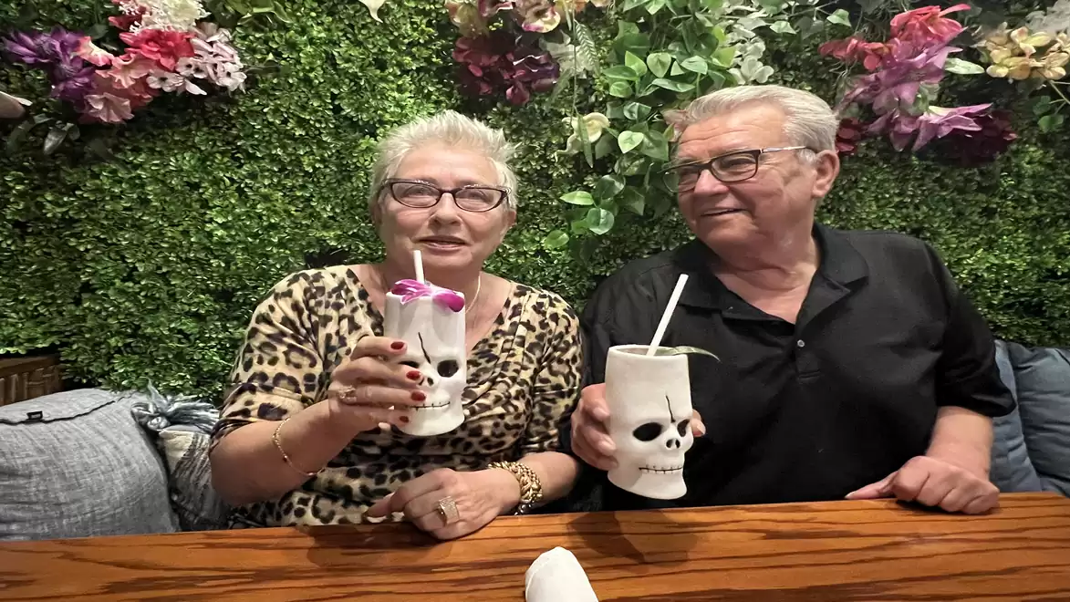 An elderly couple sits in front of a green plant wall holding colorful drinks in skull cups.