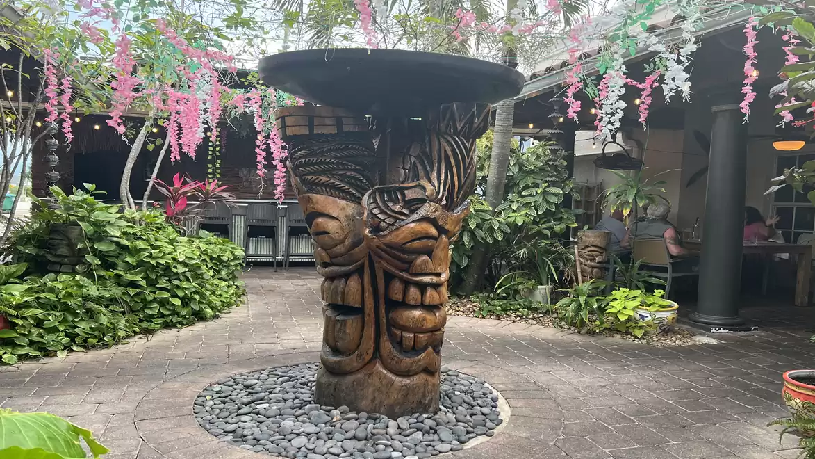 A decorative area in a garden featuring a large wooden carving artwork in the center, surrounded by plants and pink floral garlands.