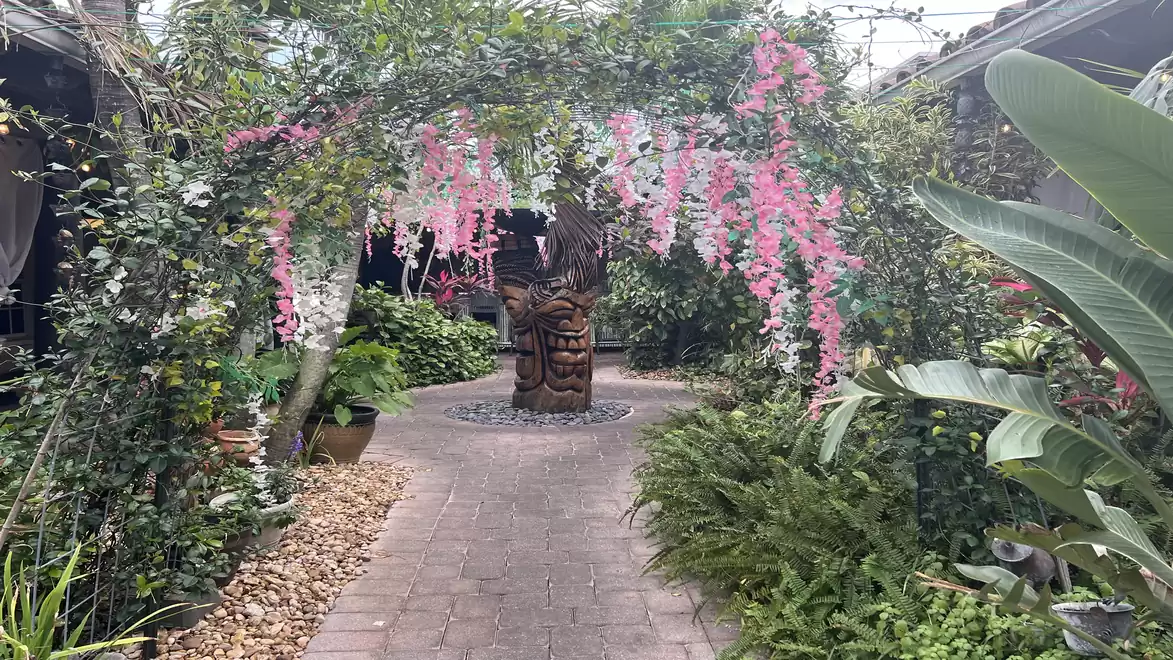An idyllic garden with a paved path, flanked by green plants and a floral vine canopy. A wooden sculpture stands in the center.