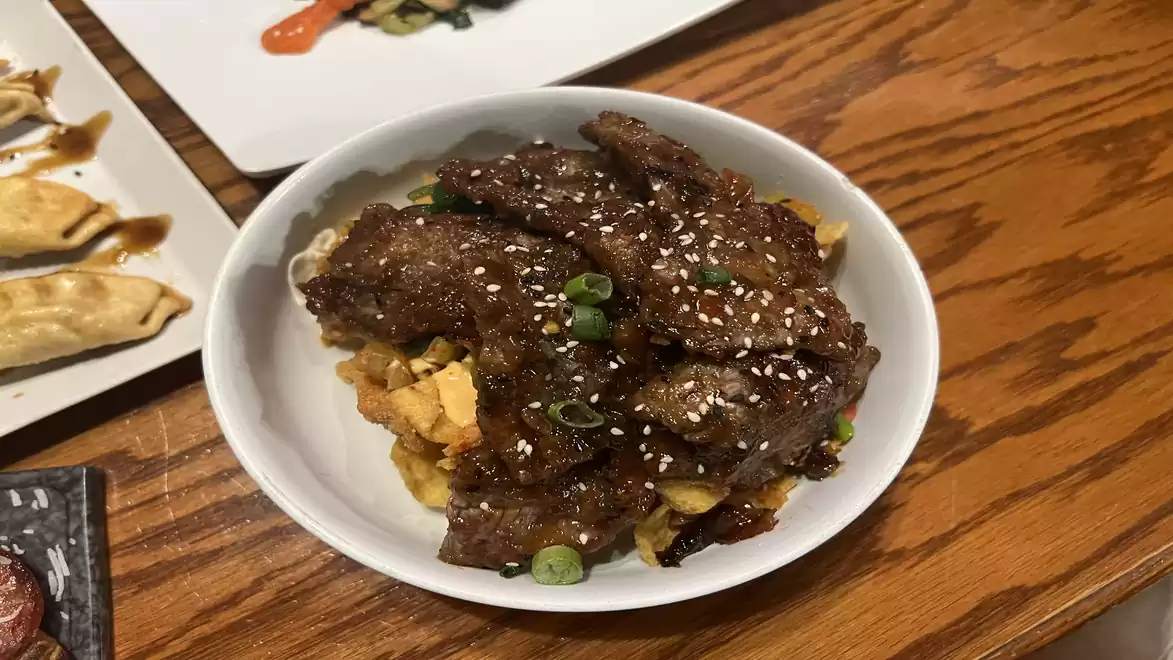 A plate of marinated meat with sesame seeds served on a wooden surface, alongside some gyoza on a rectangular plate.