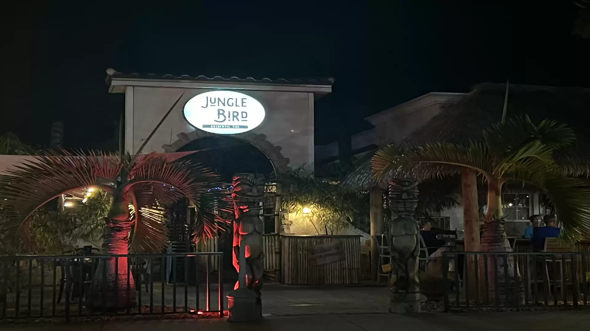 Night shot of a restaurant named Jungle Bird, illuminated by gentle lights, with palm trees in the foreground.