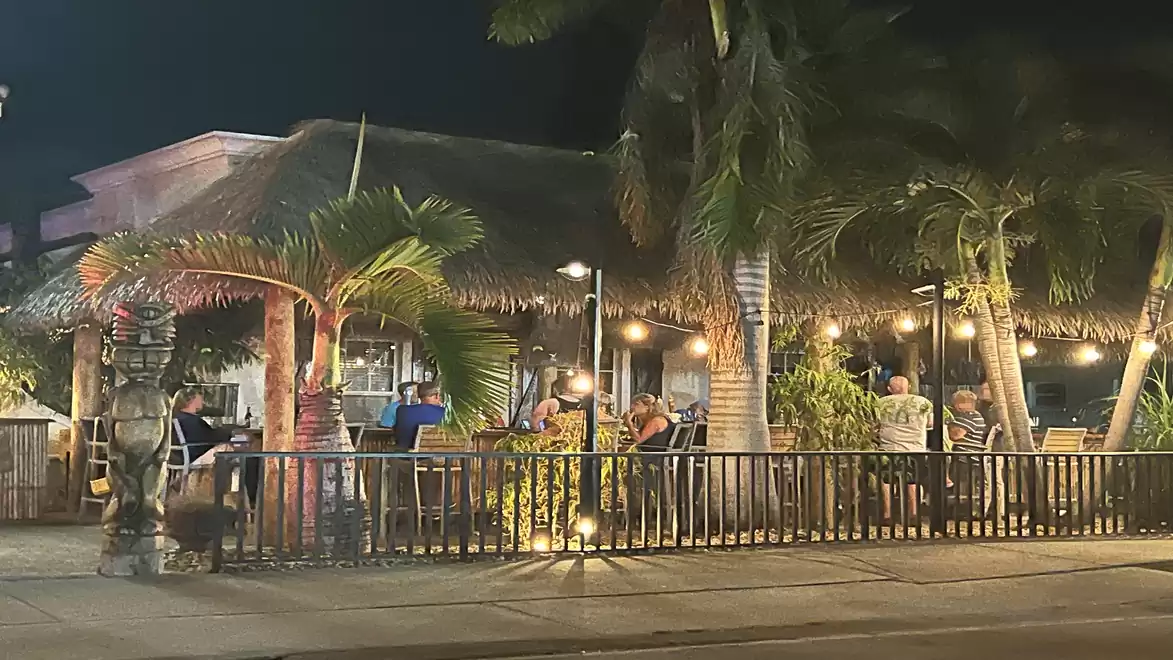 Night view of a cozy restaurant with palm trees and a thatched roof. People are sitting outside enjoying their meals under ambient lighting.