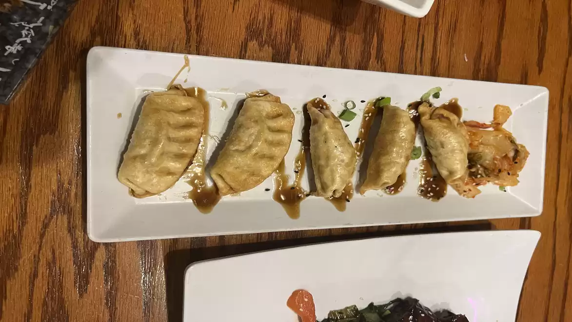 A rectangular white plate with seven golden pan-fried dumplings, served on a wooden table. The dumplings are drizzled with a brown sauce.