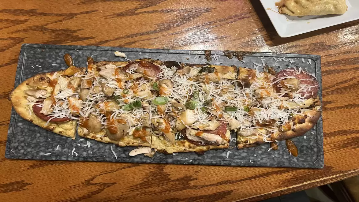 A rectangular pizza with various toppings like chicken, cheese, and vegetables, served on a black stone platter.
