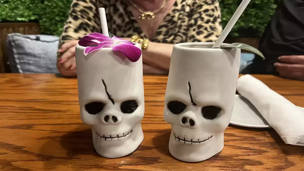 Two skull mugs with straws on a wooden table, one decorated with a purple flower, a person in a leopard print top in the background.