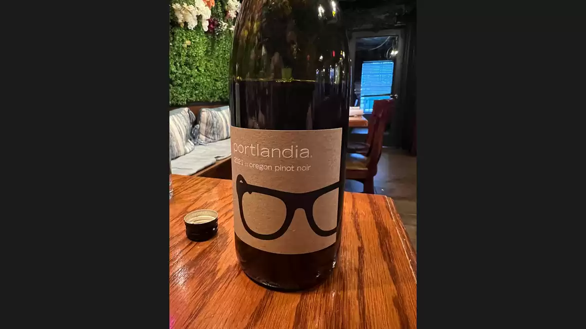Wine bottle with 'Portlandia Vintner's Pinot Noir' label featuring glasses design on wooden table in cozy restaurant.