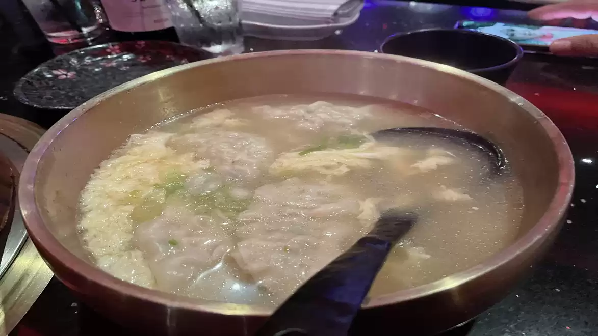 A bowl of hot soup with wontons and vegetables, served on a table with a spoon.