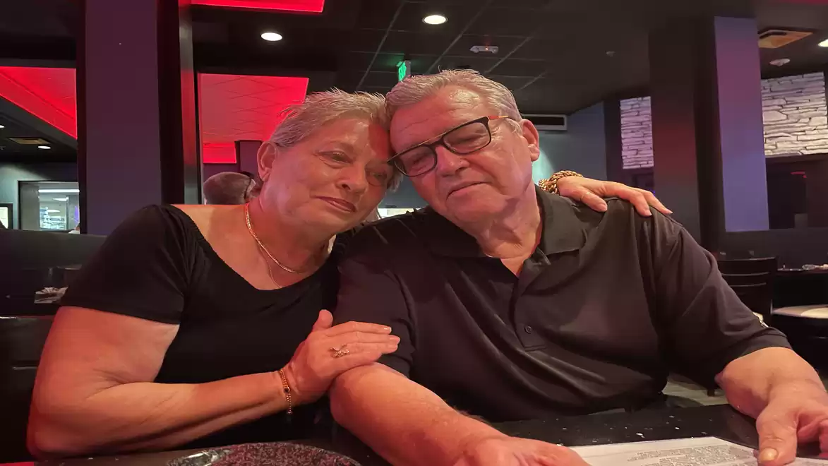 An elderly couple sits in a dimly lit restaurant, enjoying their time together. The woman rests her head on the man's shoulder, who is wearing glasses.