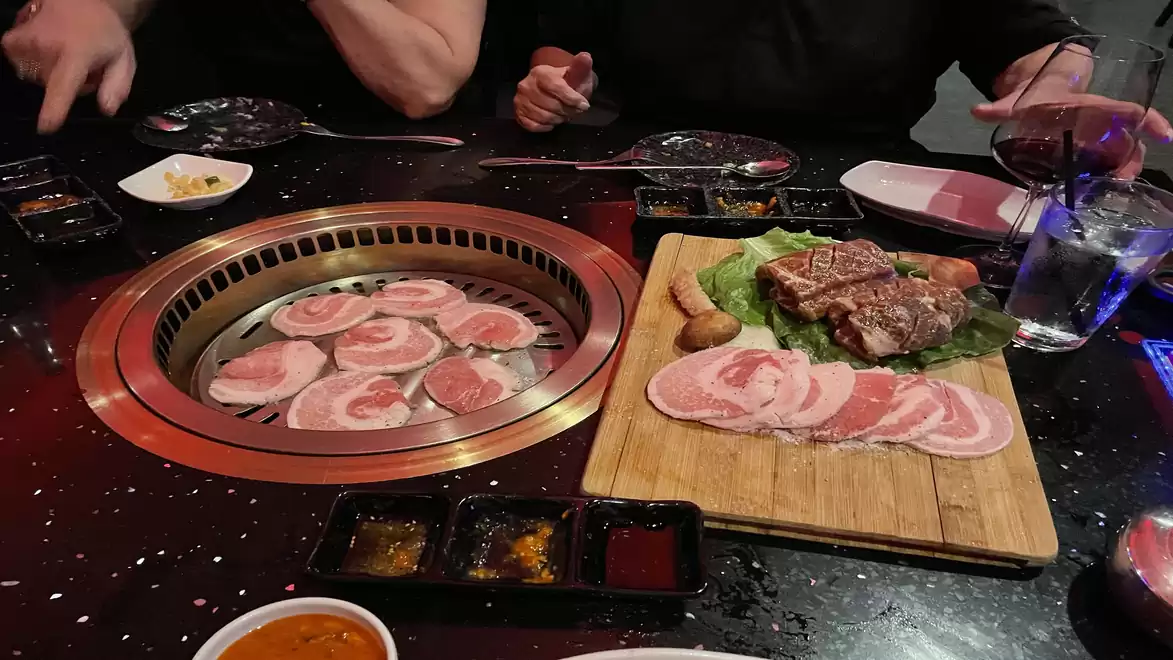 Korean BBQ table with raw meat on a wooden board and a grill with browning meat slices, surrounded by sauce dishes and plates.