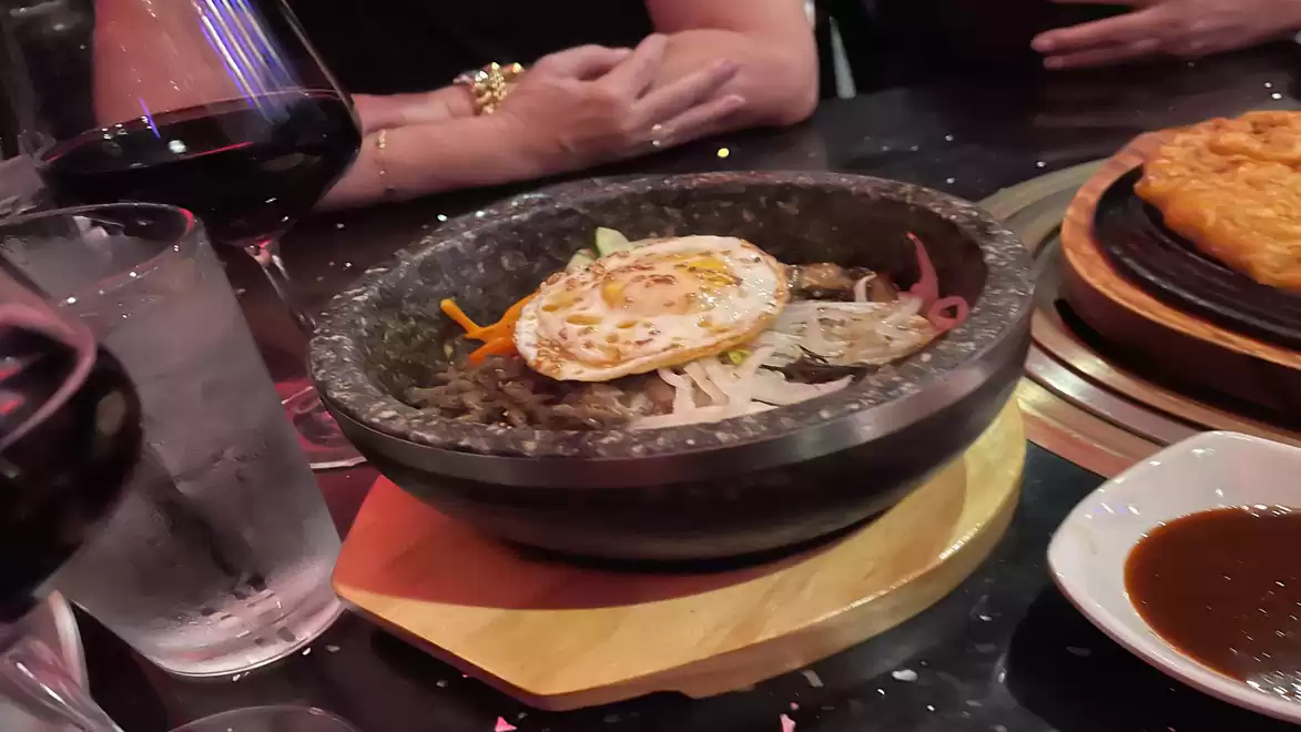 A Korean dish with an egg served in a hot stone bowl, accompanied by a glass of red wine and a bowl of sauce on a table.
