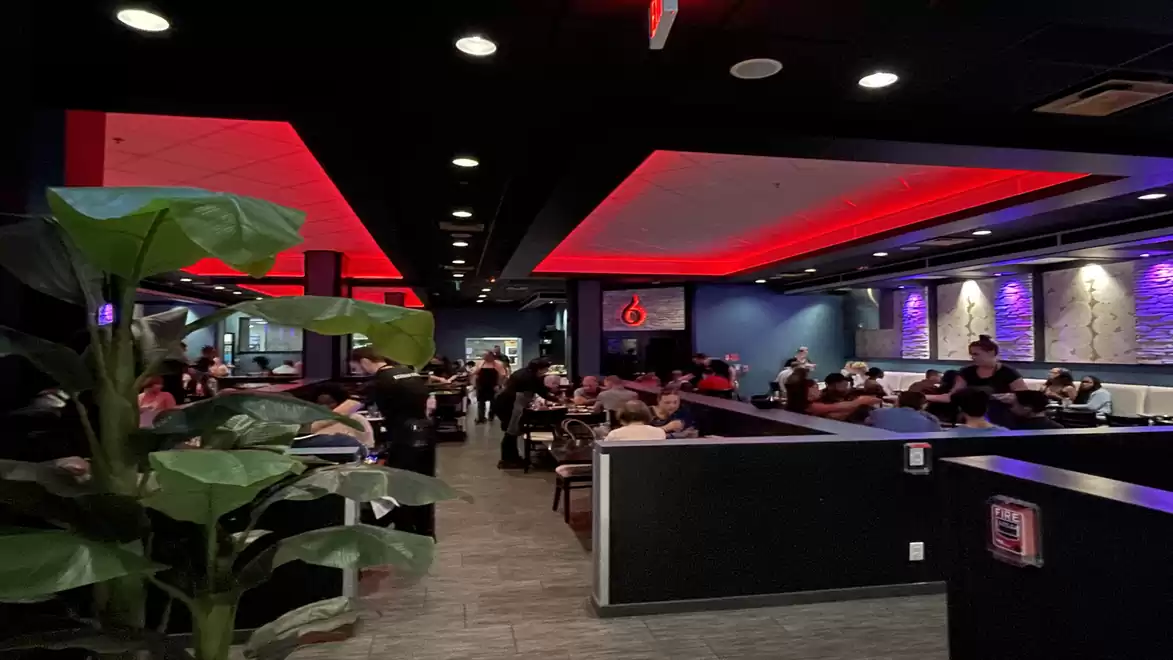 A modern restaurant with red ceiling lighting and many guests seated at tables. A large plant stands in the foreground.